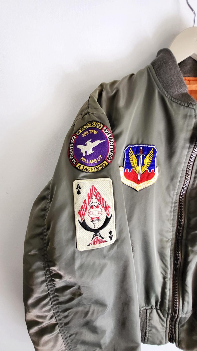 Puede incluir: Chaqueta bomber verde oliva con m&uacute;ltiples parches bordados. Los parches incluyen un avi&oacute;n morado y blanco, un escudo con una espada y alas, y un dise&ntilde;o de carta de juego con el texto "100 MISSIONS". La chaqueta tiene cuello y pu&ntilde;os acanalados.