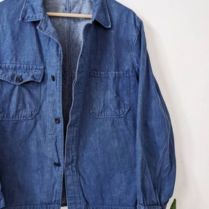 May include: A blue denim work shirt with a button-up front and two chest pockets. The shirt is hanging on a wooden hanger.