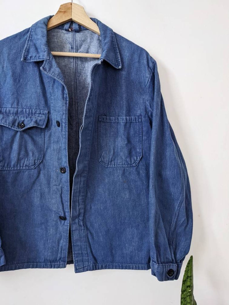 May include: A blue denim work shirt with a button-up front and two chest pockets. The shirt is hanging on a wooden hanger.