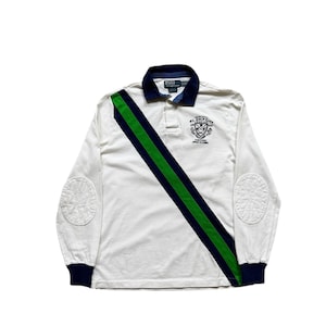 May include: White long-sleeve rugby shirt with a navy blue collar and cuffs. A diagonal green and navy stripe crosses the front. Features elbow patches and a crest graphic on the chest. The shirt has the Polo Ralph Lauren logo.