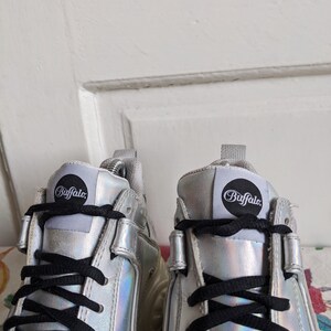 Op de afbeelding: Een paar zilveren platform sneakers met zwarte veters. De sneakers hebben een rond logo aan de zijkant met de tekst "Buffalo".