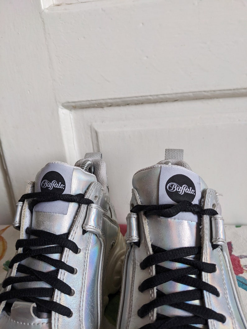 Op de afbeelding: Een paar zilveren platform sneakers met zwarte veters. De sneakers hebben een rond logo aan de zijkant met de tekst "Buffalo".