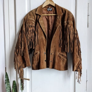 May include: Brown suede jacket with fringe detailing. The jacket features a classic collar, button closure, and pockets. The fringe is along the sleeves and chest. The jacket is hanging on a wooden hanger.
