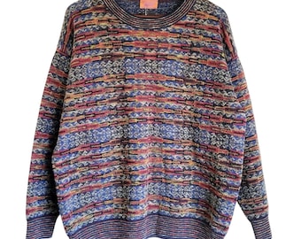 May include: A multi-colored sweater with a crew neck and long sleeves. The sweater features a pattern of horizontal stripes and geometric shapes in shades of blue, red, orange, and yellow. The sweater is hanging on a wooden hanger.