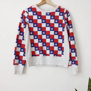 Może przedstawiać: Biała bluza z długim rękawem w czerwono-niebieską kratę z powtarzającym się logo Champion.