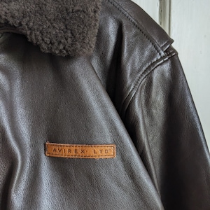 May include: Close-up of a brown leather jacket with a shearling collar. The jacket features a leather patch with the text "AVIREX LTD" in a rectangular shape. The jacket has a classic design.