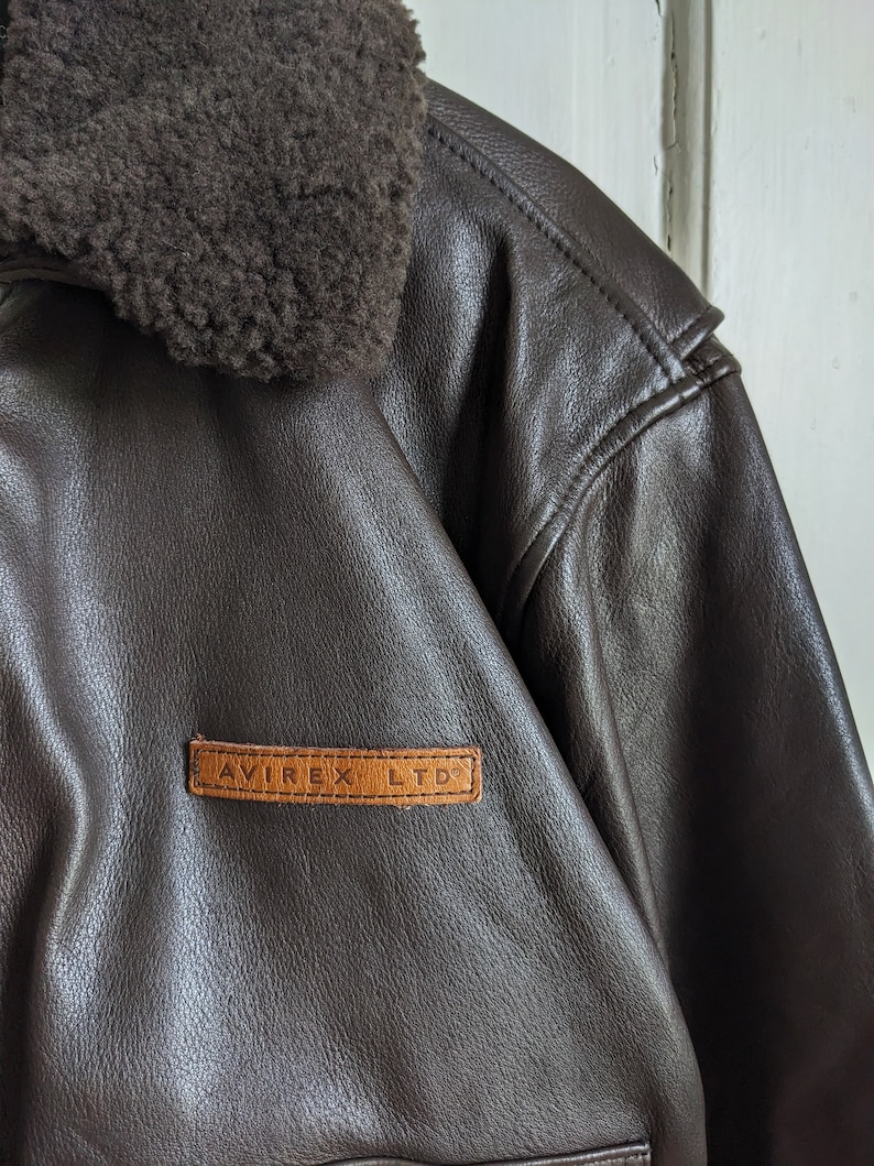 May include: Close-up of a brown leather jacket with a shearling collar. The jacket features a leather patch with the text "AVIREX LTD" in a rectangular shape. The jacket has a classic design.