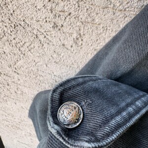 Puede incluir: Primer plano de un pu&ntilde;o de chaqueta vaquera gris oscuro con un bot&oacute;n plateado. El bot&oacute;n tiene un dise&ntilde;o en relieve detallado. La tela vaquera muestra textura y costuras visibles.