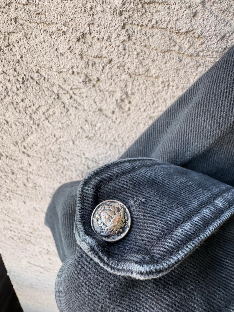 Puede incluir: Primer plano de un pu&ntilde;o de chaqueta vaquera gris oscuro con un bot&oacute;n plateado. El bot&oacute;n tiene un dise&ntilde;o en relieve detallado. La tela vaquera muestra textura y costuras visibles.