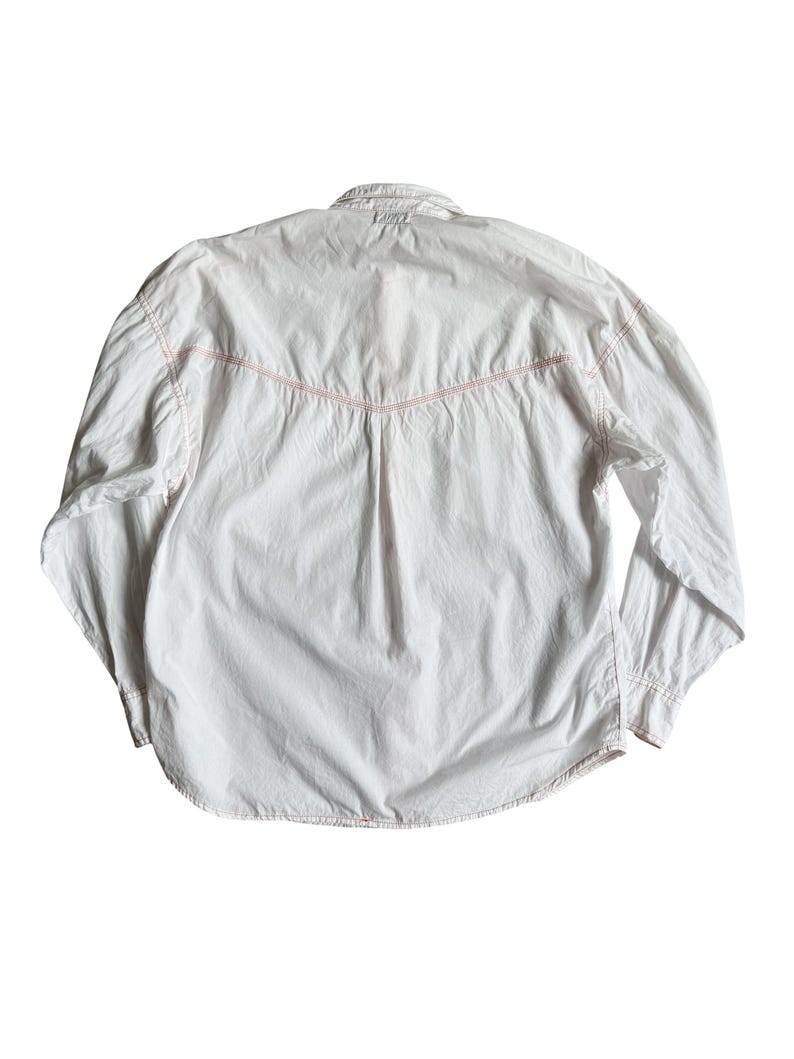 May include: A white long-sleeved button-down shirt featuring a pointed western-style back yoke with contrasting orange stitching along the seams. The shirt has a standard collar, long sleeves with buttoned cuffs, and a slightly curved hemline.