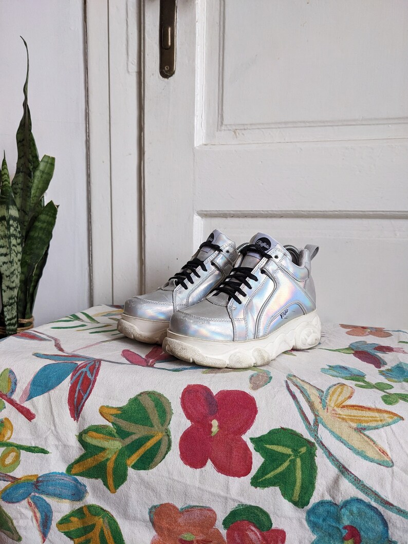 Op de afbeelding: Een paar zilveren platform sneakers met zwarte veters. De sneakers hebben een dikke zool en staan op een stoffen ondergrond met bloemenprint.