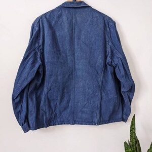 May include: A dark blue denim jacket with a button-up front and long sleeves. The jacket is hanging on a wooden hanger against a white wall.