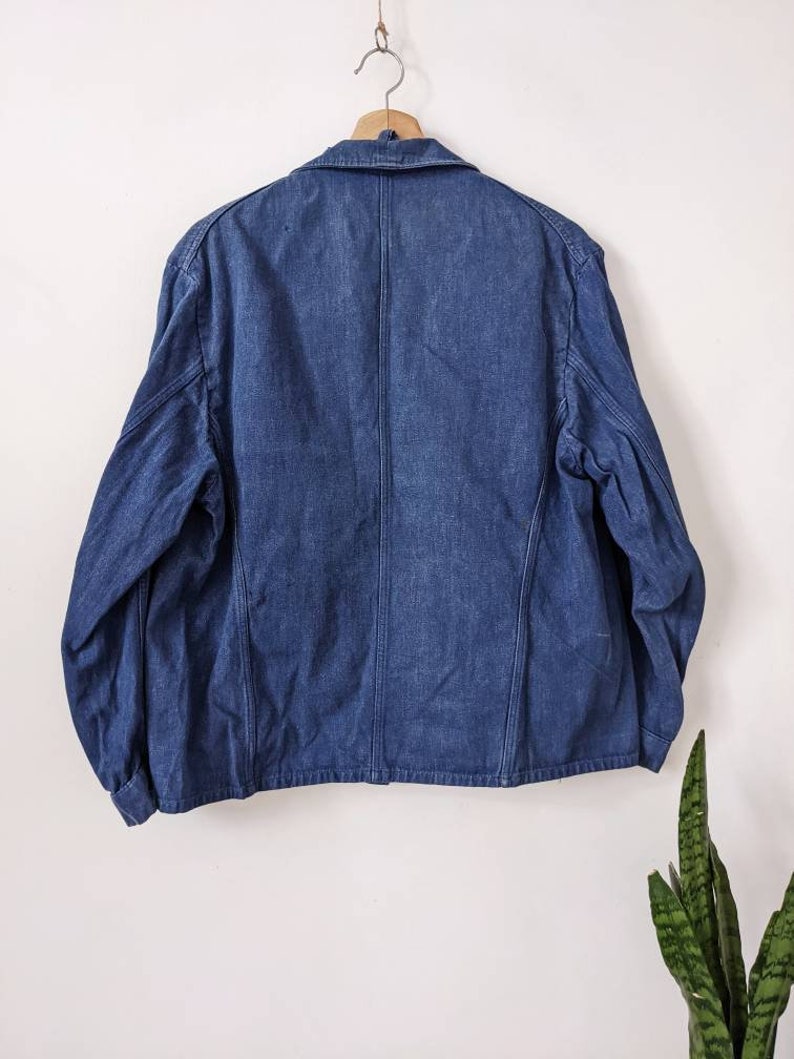 May include: A dark blue denim jacket with a button-up front and long sleeves. The jacket is hanging on a wooden hanger against a white wall.
