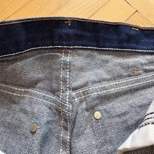 Op de afbeelding: Close-up van de tailleband en zakken van een blauwe denim jeans. De jeans heeft witte stiksels, metalen klinknagels en een knoopsluiting. De denim stof heeft een getextureerd uiterlijk.