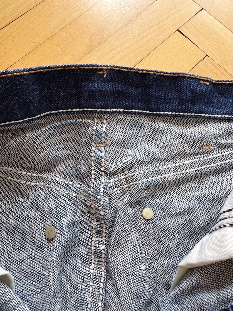Op de afbeelding: Close-up van de tailleband en zakken van een blauwe denim jeans. De jeans heeft witte stiksels, metalen klinknagels en een knoopsluiting. De denim stof heeft een getextureerd uiterlijk.