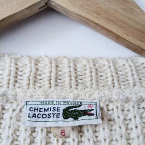 May include: A white knitted sweater with a Lacoste logo tag. The tag reads "Chemise Lacoste" and "Made in France".