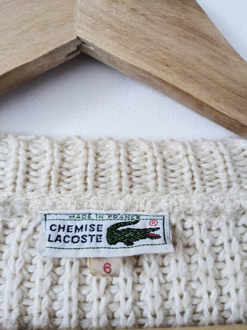 May include: A white knitted sweater with a Lacoste logo tag. The tag reads "Chemise Lacoste" and "Made in France".