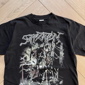 May include: Black t-shirt with the band name "Suffocation" in a stylized font above a graphic of skeletons and a graveyard scene. The text "...Evil lies, death denied" is below the graphic.