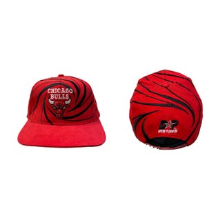 Puede incluir: Gorra de béisbol roja y negra con un logotipo de los Chicago Bulls y un patrón de espiral. La parte posterior de la gorra tiene una estrella negra y el texto "Drew Pearson".