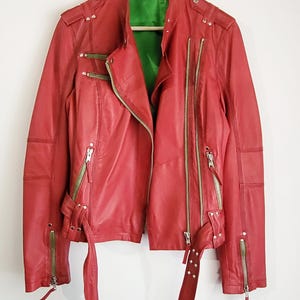 May include: A red leather jacket with multiple zippers and a green interior lining. The jacket has a belted waist and is hanging on a hanger. The brand name "MaxMara" is visible on the hanger.