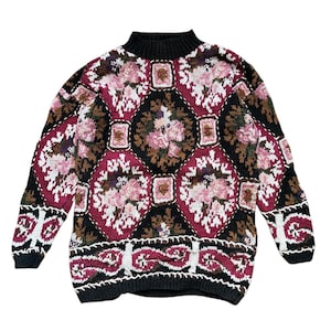 Op de afbeelding: Een vintage trui met een hoge halslijn. De trui heeft een zwarte basis met een herhalend patroon van bloemmotieven in roze, wit, bruin en bordeauxrood. De manchetten en zoom zijn zwart en het ontwerp bevat witte stiksels.