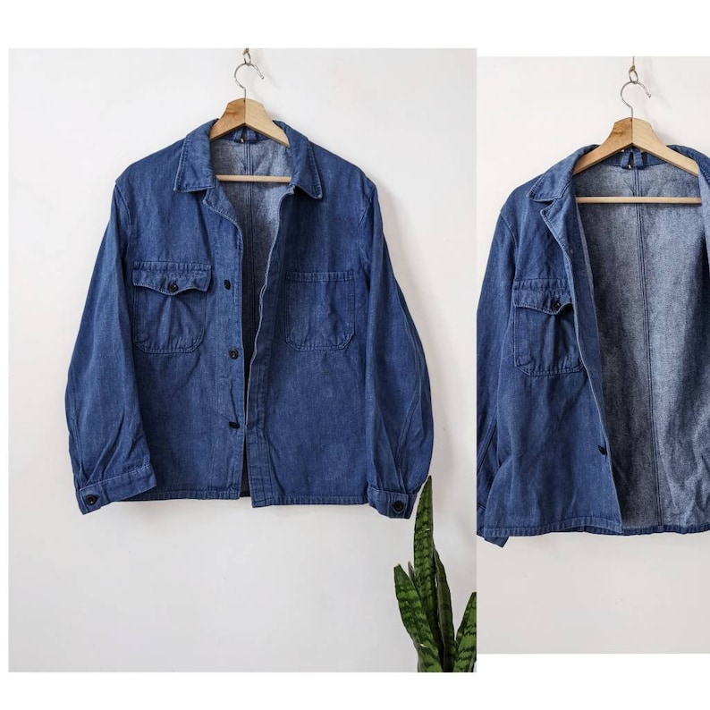 May include: A blue denim work shirt with two chest pockets and a button-up front. The shirt is hanging on a wooden hanger.