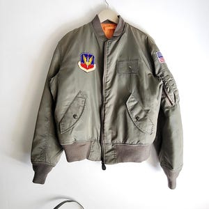 Puede incluir: Chaqueta bomber verde oliva con cuello y pu&ntilde;os marrones. Cuenta con un parche colorido en el pecho y un peque&ntilde;o parche de la bandera estadounidense en la manga. La chaqueta tiene cierre de cremallera y dos bolsillos delanteros.