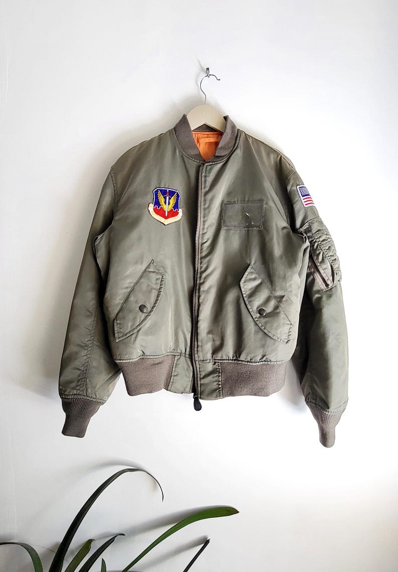 Puede incluir: Chaqueta bomber verde oliva con cuello y pu&ntilde;os marrones. Cuenta con un parche colorido en el pecho y un peque&ntilde;o parche de la bandera estadounidense en la manga. La chaqueta tiene cierre de cremallera y dos bolsillos delanteros.