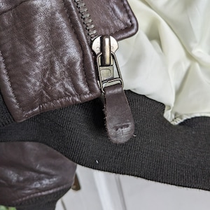 May include: Close-up of a brown leather jacket with a metal zipper. The jacket has a quilted design on the upper portion and a dark brown ribbed cuff. The interior lining is visible, with text that reads "NAVAL AIR BASIC".