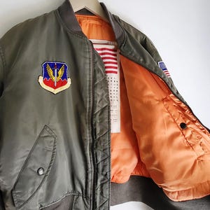 Puede incluir: Chaqueta bomber verde oliva con forro naranja brillante. Cuenta con un parche con un escudo y alas, un parche de la bandera estadounidense y cuello y pu&ntilde;os acanalados. La chaqueta est&aacute; colgada en una percha de madera.