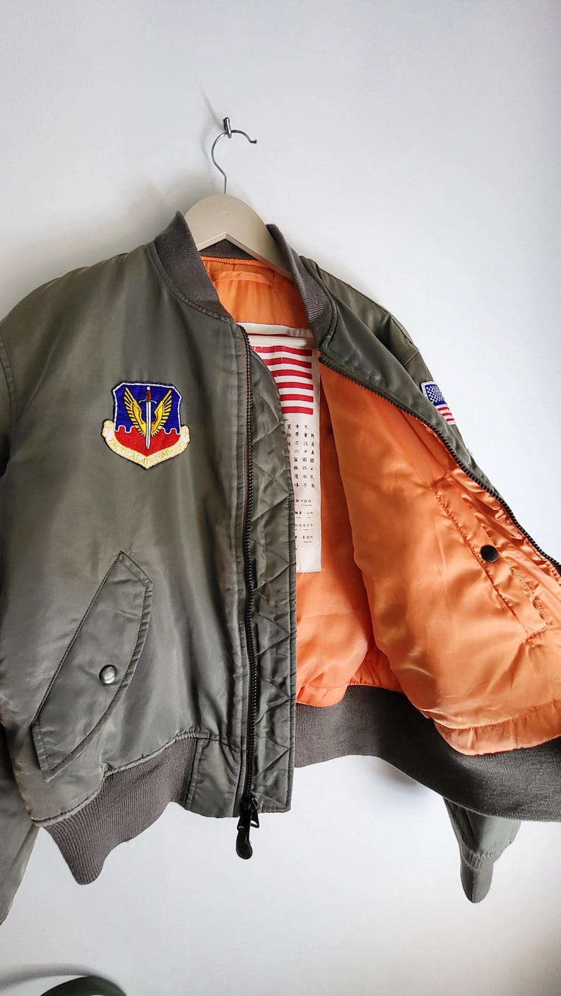 Puede incluir: Chaqueta bomber verde oliva con forro naranja brillante. Cuenta con un parche con un escudo y alas, un parche de la bandera estadounidense y cuello y pu&ntilde;os acanalados. La chaqueta est&aacute; colgada en una percha de madera.
