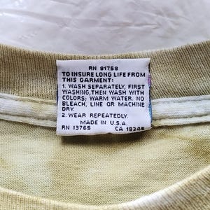 Puede incluir: Primer plano de una etiqueta en una camiseta te&ntilde;ida con tinte anudado. La etiqueta dice "RN 81758" y proporciona instrucciones de lavado: lavar por separado, luego con colores, agua tibia, sin lej&iacute;a, secar al aire o a m&aacute;quina. Tambi&eacute;n dice "Made in U.S.A."