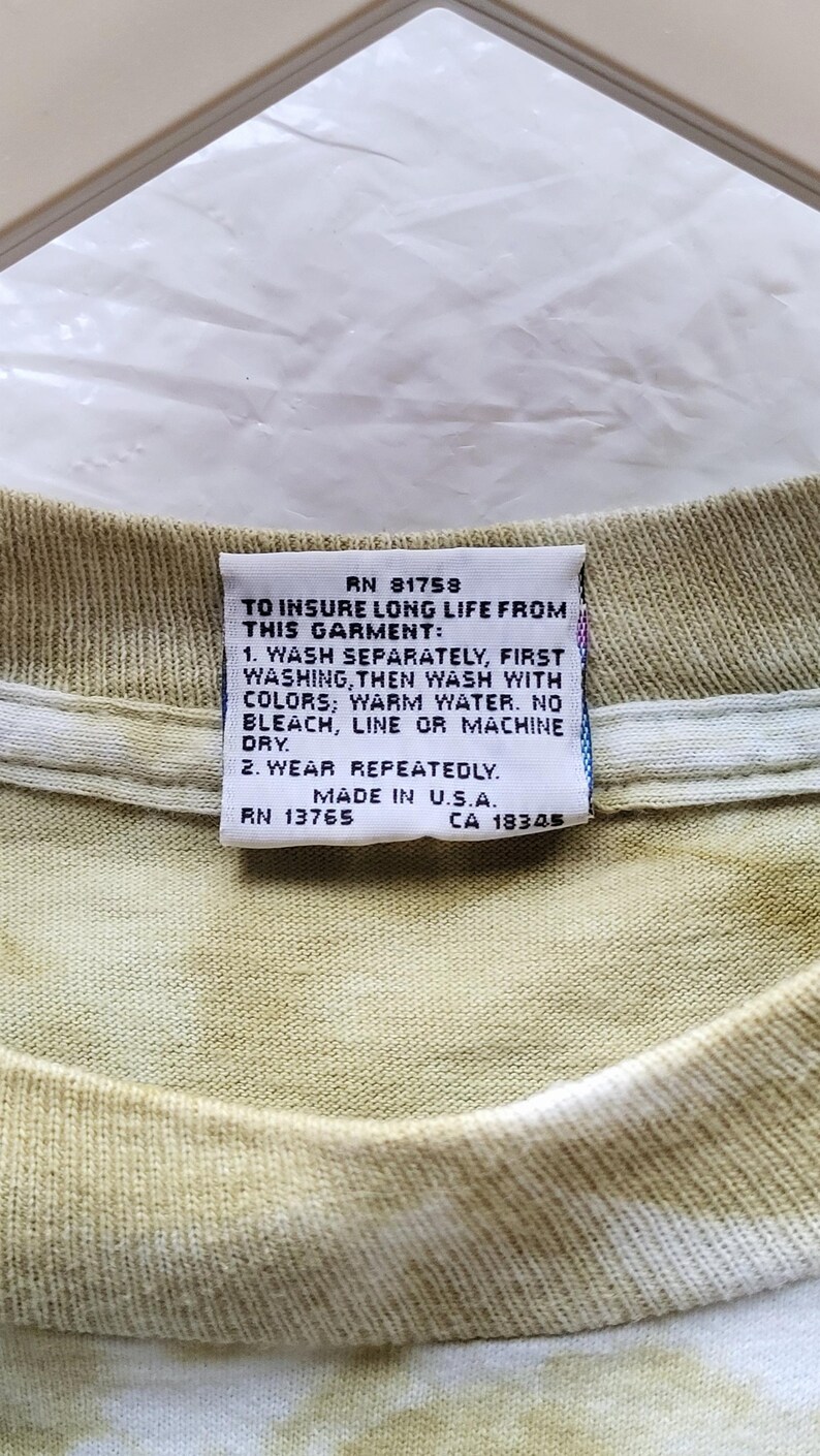 Puede incluir: Primer plano de una etiqueta en una camiseta te&ntilde;ida con tinte anudado. La etiqueta dice "RN 81758" y proporciona instrucciones de lavado: lavar por separado, luego con colores, agua tibia, sin lej&iacute;a, secar al aire o a m&aacute;quina. Tambi&eacute;n dice "Made in U.S.A."