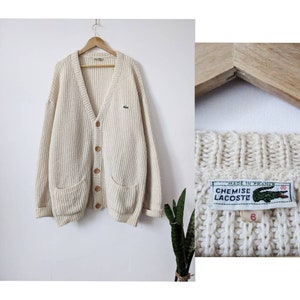 May include: A cream-colored knitted cardigan with a V-neck, button closure, and two patch pockets. The cardigan has a green crocodile logo on the left chest and a label that reads "Chemise Lacoste" and "Made in France".