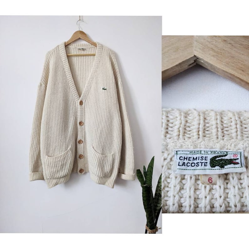 May include: A cream-colored knitted cardigan with a V-neck, button closure, and two patch pockets. The cardigan has a green crocodile logo on the left chest and a label that reads "Chemise Lacoste" and "Made in France".