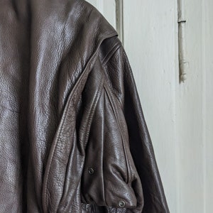 May include: Close-up of a brown leather jacket with a shearling collar. The jacket has a textured surface and a pocket on the sleeve with two metal snap closures. The jacket is hanging against a white door.