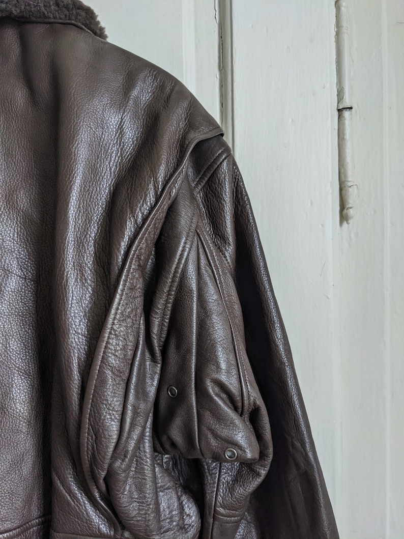 May include: Close-up of a brown leather jacket with a shearling collar. The jacket has a textured surface and a pocket on the sleeve with two metal snap closures. The jacket is hanging against a white door.