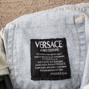 K&ouml;nnte beinhalten: Nahaufnahme eines hellblauen Jeanskleidungsst&uuml;cks mit einem schwarzen Etikett. Das Etikett zeigt "VERSACE JEANS COUTURE" in wei&szlig;er Schrift. Zus&auml;tzlicher Text auf Italienisch ist ebenfalls sichtbar.