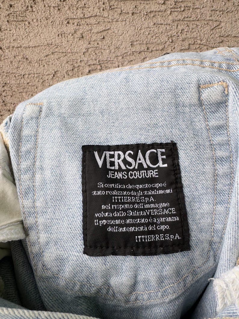 K&ouml;nnte beinhalten: Nahaufnahme eines hellblauen Jeanskleidungsst&uuml;cks mit einem schwarzen Etikett. Das Etikett zeigt "VERSACE JEANS COUTURE" in wei&szlig;er Schrift. Zus&auml;tzlicher Text auf Italienisch ist ebenfalls sichtbar.