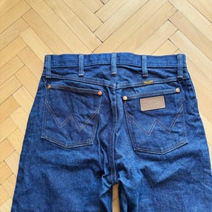 Op de afbeelding: Donkerblauwe denim jeans met het Wrangler-logo op de achterzak. De jeans hebben koperen klinknagels en een klassiek ontwerp met vijf zakken. De stiksels zijn in een contrasterende oranje kleur.