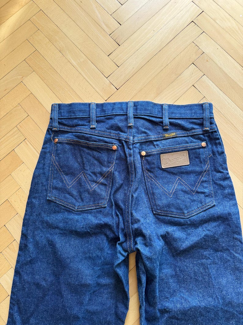Op de afbeelding: Donkerblauwe denim jeans met het Wrangler-logo op de achterzak. De jeans hebben koperen klinknagels en een klassiek ontwerp met vijf zakken. De stiksels zijn in een contrasterende oranje kleur.