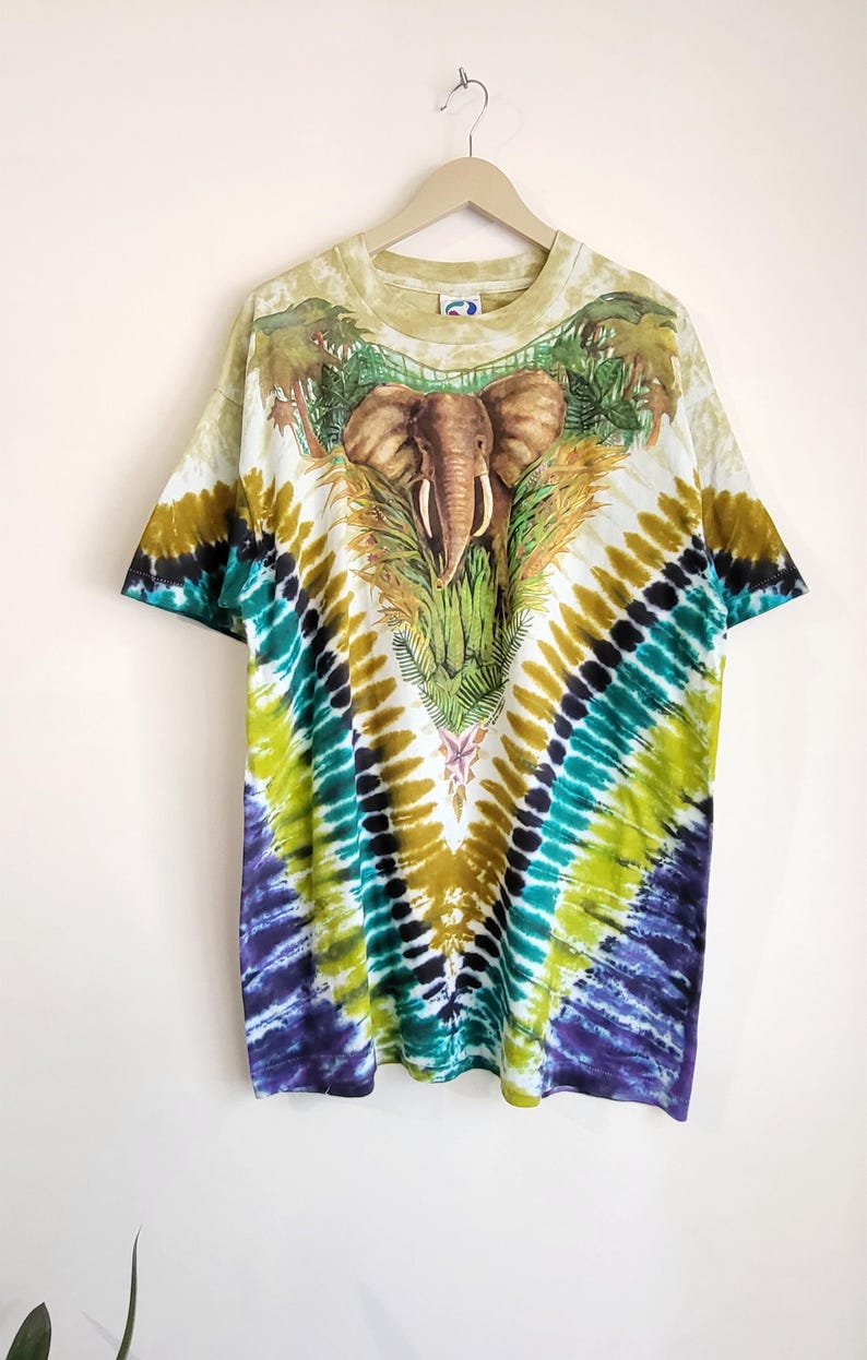 Puede incluir: Una camiseta te&ntilde;ida con un dise&ntilde;o de elefante. La camiseta tiene una base blanca con un colorido patr&oacute;n tie-dye en tonos verdes, azules y amarillos. El elefante est&aacute; representado con un dise&ntilde;o detallado, rodeado de follaje tropical.