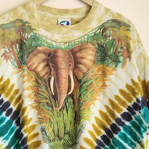 Puede incluir: Una camiseta te&ntilde;ida con un dise&ntilde;o de elefante rodeado de follaje tropical. La camiseta presenta un patr&oacute;n tie-dye colorido en tonos verdes, amarillos, azules y negros. La prenda est&aacute; colgada de una percha de madera.