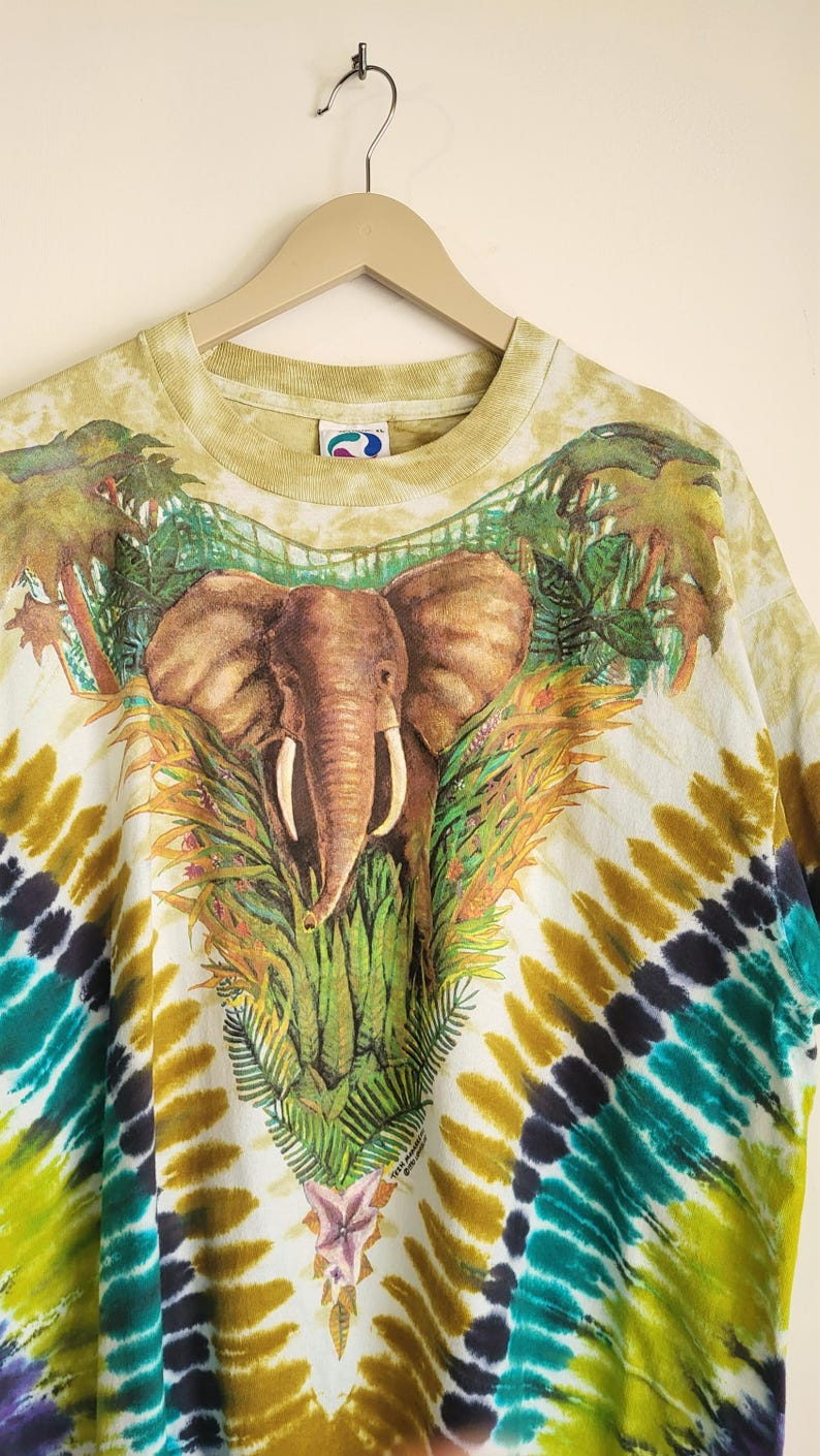 Puede incluir: Una camiseta te&ntilde;ida con un dise&ntilde;o de elefante rodeado de follaje tropical. La camiseta presenta un patr&oacute;n tie-dye colorido en tonos verdes, amarillos, azules y negros. La prenda est&aacute; colgada de una percha de madera.