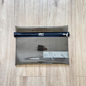 May include: A rectangular, translucent dark gray pouch made of a flexible material. It features a black horizontal zipper closure across the top with a silver circular pull tab. The text MM6 Maison Margiela is printed in white on the zipper strip.