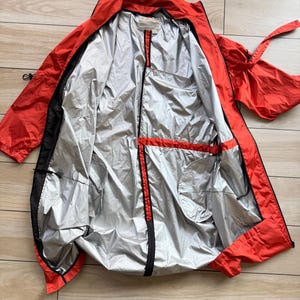 Puede incluir: Chaqueta naranja abierta con forro plateado, ribete negro y cremallera completa. La chaqueta tiene dos bolsillos delanteros y un cordón en la cintura. La chaqueta está tendida sobre una superficie de madera.