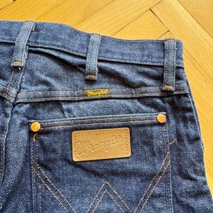 Op de afbeelding: Close-up van de achterkant van een donkerblauwe denim jeans. De jeans heeft een bruin leren label met de merknaam "Wrangler" en een klein geel label met dezelfde merknaam. De stiksels zijn oranje.