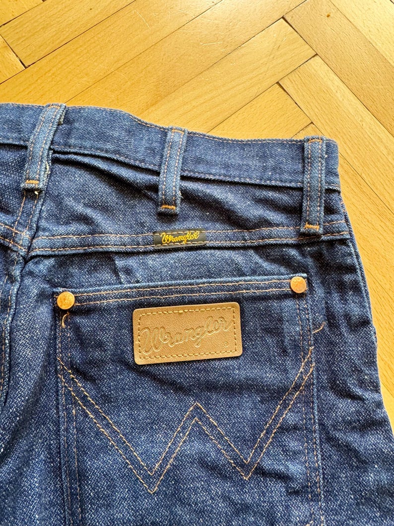 Op de afbeelding: Close-up van de achterkant van een donkerblauwe denim jeans. De jeans heeft een bruin leren label met de merknaam "Wrangler" en een klein geel label met dezelfde merknaam. De stiksels zijn oranje.