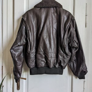 May include: A brown leather bomber jacket with a dark brown faux fur collar. The jacket has a ribbed black hem and cuffs. The back of the jacket is visible, showing the textured leather and stitching details. The jacket is hanging on a metal hanger.
