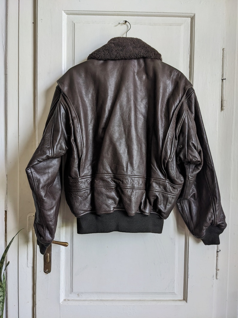May include: A brown leather bomber jacket with a dark brown faux fur collar. The jacket has a ribbed black hem and cuffs. The back of the jacket is visible, showing the textured leather and stitching details. The jacket is hanging on a metal hanger.